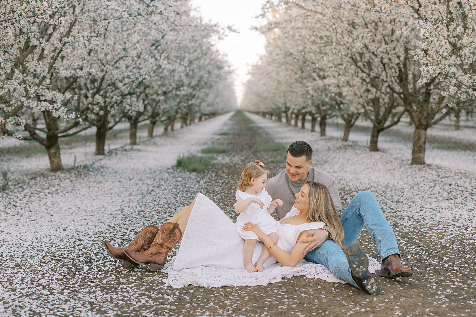 A family of three posing in a Stockton almond blossom orchard during golden hour, with soft pastel tones and warm, natural light enhancing the serene beauty of the blooms