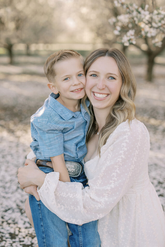 A mother and her young son share a joyful embrace in an almond orchard during peak bloom. Captured in soft golden-hour light by Sacramento family photographer Nicole Hodgson.