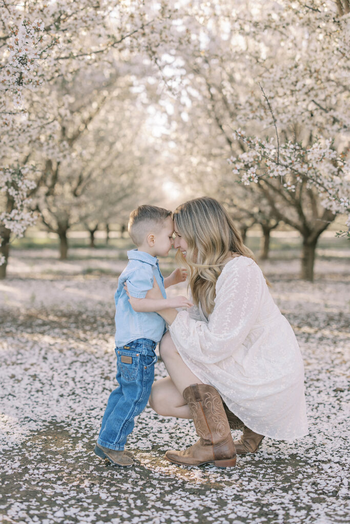 A mother and her young son share a sweet forehead-to-forehead moment in an almond orchard, surrounded by soft white blossoms. Captured by Sacramento family photographer Nicole Hodgson.