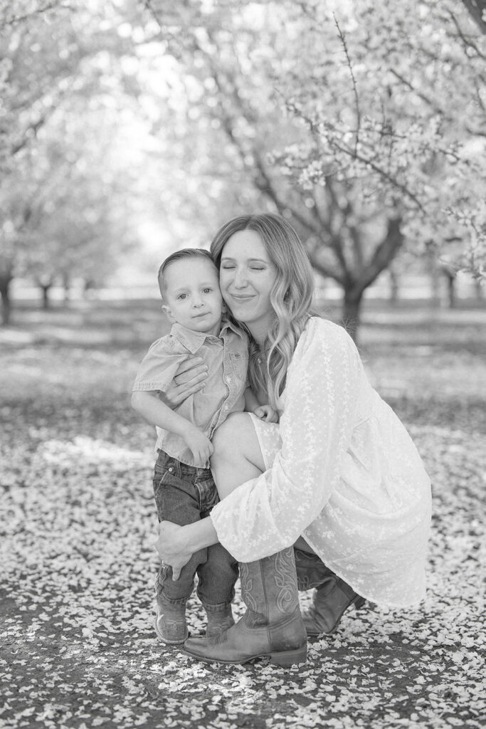 A mother kneels beside her young son in an almond orchard, sharing a quiet moment among the blossoms. Captured in black and white by Sacramento family photographer Nicole Hodgson.