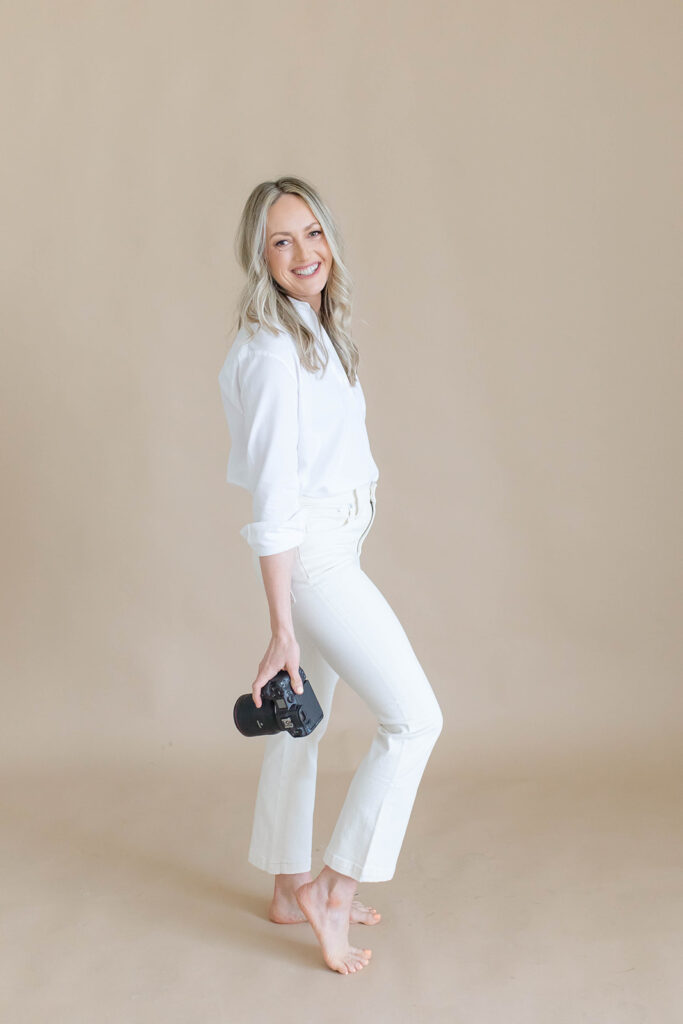 Nicole Hodgson, a Sacramento family photographer, smiles while holding her camera in a light-filled studio. She specializes in natural, timeless family photography.